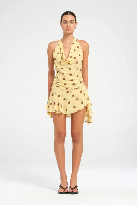 Yellow - A woman wearing a pale yellow halter neck mini dress with large brown polka dots, featuring a cowl neckline, side ruching, and an asymmetrical ruffled hem.
