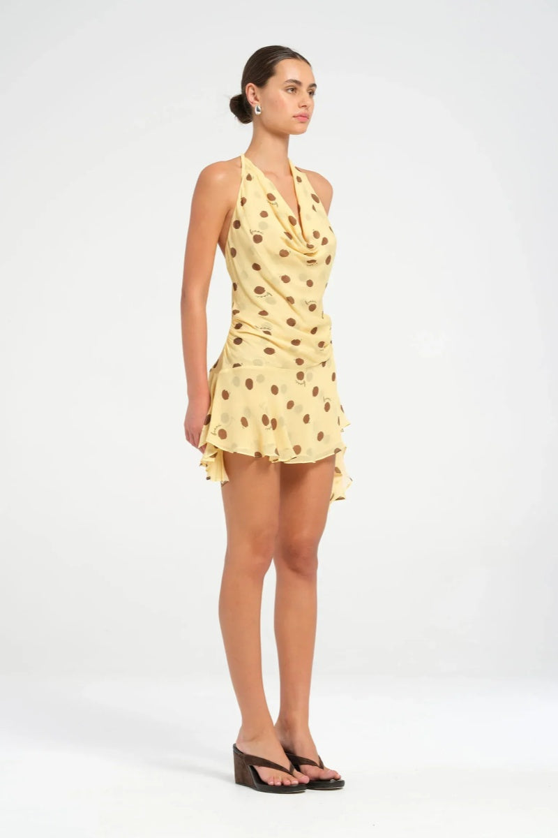 Yellow - A woman wearing a pale yellow halter neck mini dress with large brown polka dots, featuring a cowl neckline, side ruching, and an asymmetrical ruffled hem.