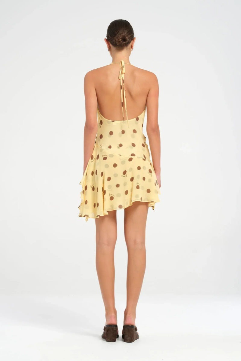 Yellow - A woman wearing a pale yellow halter neck mini dress with large brown polka dots, featuring a cowl neckline, side ruching, and an asymmetrical ruffled hem.