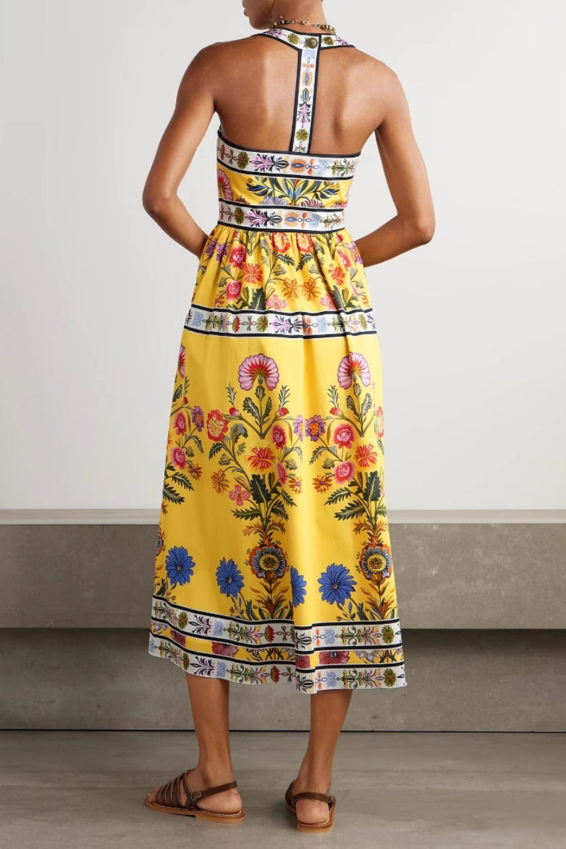 Woman wearing a bright yellow halter midi dress featuring a vibrant botanical floral print and contrasting geometric borders, styled with a woven tote bag and brown sandals.