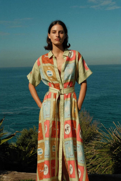Multicolored - A woman wearing a long, short-sleeved shirt dress featuring a colourful patchwork tile print with whimsical motifs. The dress has a button-down front, a collar, a matching waist belt, and side pockets.