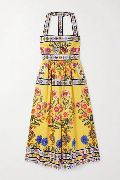 Yellow - Woman wearing a bright yellow halter midi dress featuring a vibrant botanical floral print and contrasting geometric borders, styled with a woven tote bag and brown sandals.