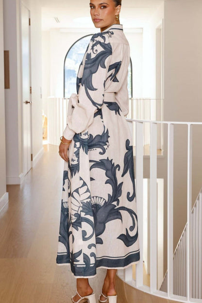 Black - Full-length view of a woman wearing a pink maxi shirt dress featuring a bold blue baroque print, long blouson sleeves, a collared neck, and a matching tie-belt at the waist.