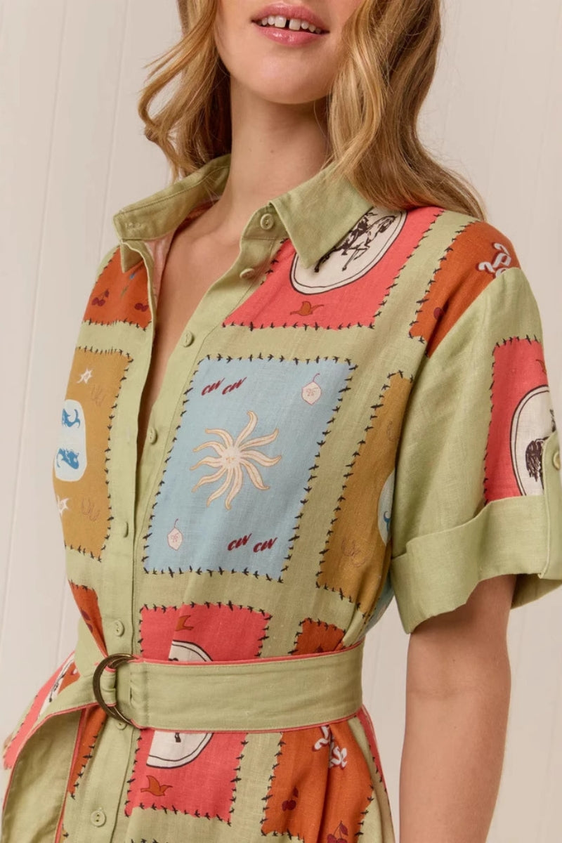 Multicolored - A woman wearing a long, short-sleeved shirt dress featuring a colourful patchwork tile print with whimsical motifs. The dress has a button-down front, a collar, a matching waist belt, and side pockets.