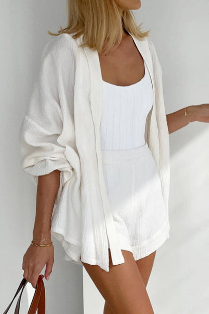 White - Woman wearing a white, crinkled-texture wrap playsuit with voluminous sleeves and a tie waist, holding a brown clutch bag.