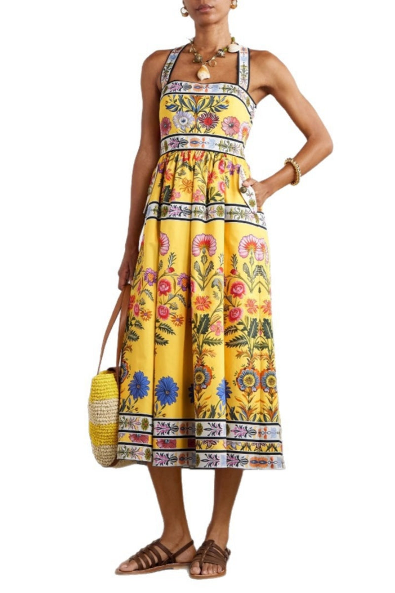Yellow - Woman wearing a bright yellow halter midi dress featuring a vibrant botanical floral print and contrasting geometric borders, styled with a woven tote bag and brown sandals.