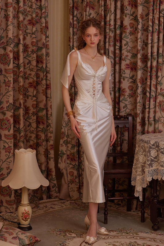 White - A woman wearing an elegant white satin midi dress featuring a corset-style lace-up bodice, ruched skirt detailing, and tie-shoulder straps, standing in a vintage-inspired room.