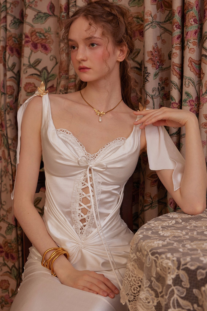 White - A woman wearing an elegant white satin midi dress featuring a corset-style lace-up bodice, ruched skirt detailing, and tie-shoulder straps, standing in a vintage-inspired room.