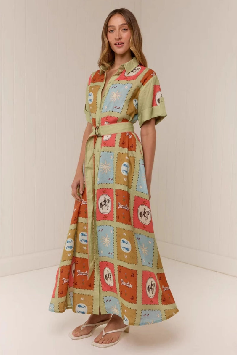 Multicolored - A woman wearing a long, short-sleeved shirt dress featuring a colourful patchwork tile print with whimsical motifs. The dress has a button-down front, a collar, a matching waist belt, and side pockets.