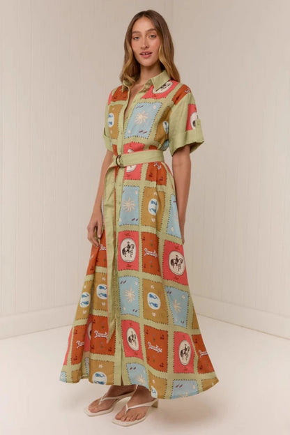 Multicolored - A woman wearing a long, short-sleeved shirt dress featuring a colourful patchwork tile print with whimsical motifs. The dress has a button-down front, a collar, a matching waist belt, and side pockets.