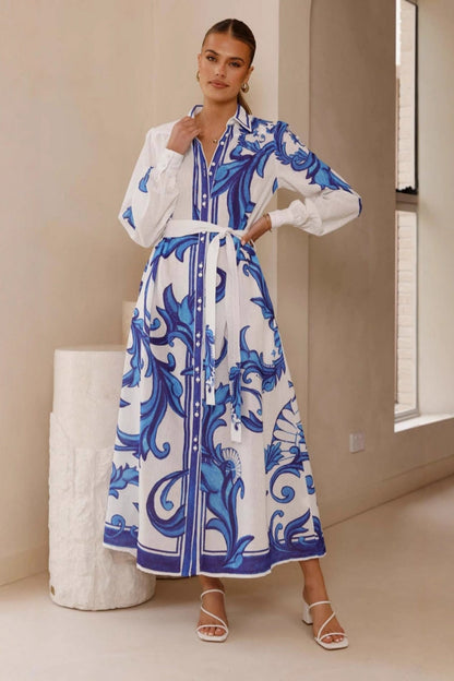 Blue - Full-length view of a woman wearing a pink maxi shirt dress featuring a bold blue baroque print, long blouson sleeves, a collared neck, and a matching tie-belt at the waist.