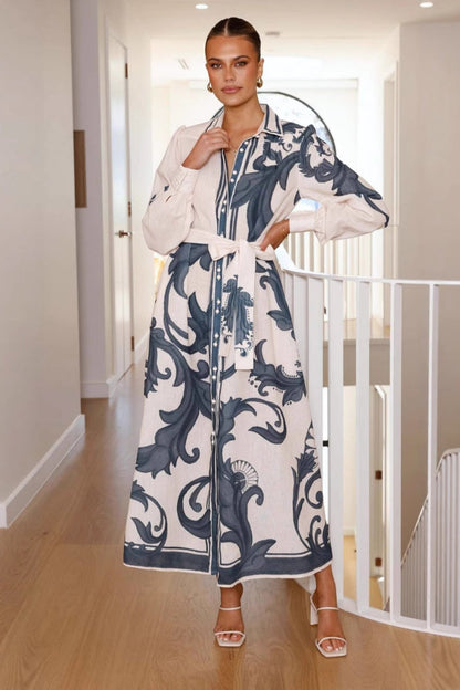 Black - Full-length view of a woman wearing a pink maxi shirt dress featuring a bold blue baroque print, long blouson sleeves, a collared neck, and a matching tie-belt at the waist.