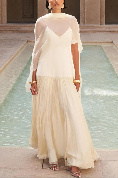 A woman wearing an elegant drop-waist maxi dress featuring a white V-neck slip bodice and a flowing, semi-sheer cream gathered skirt, styled with a matching neck scarf.