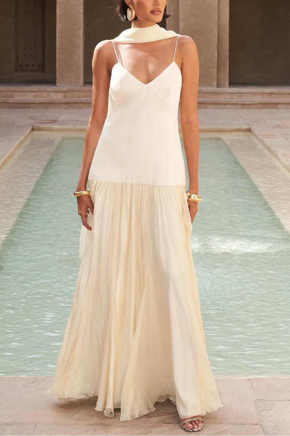 A woman wearing an elegant drop-waist maxi dress featuring a white V-neck slip bodice and a flowing, semi-sheer cream gathered skirt, styled with a matching neck scarf.