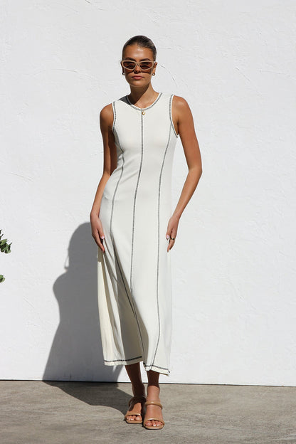 A woman stands against a white wall wearing a sleeveless, form-fitting, off-white midi dress. The dress has a high neckline and is detailed with dark vertical contrast stitching along the seams and armholes.