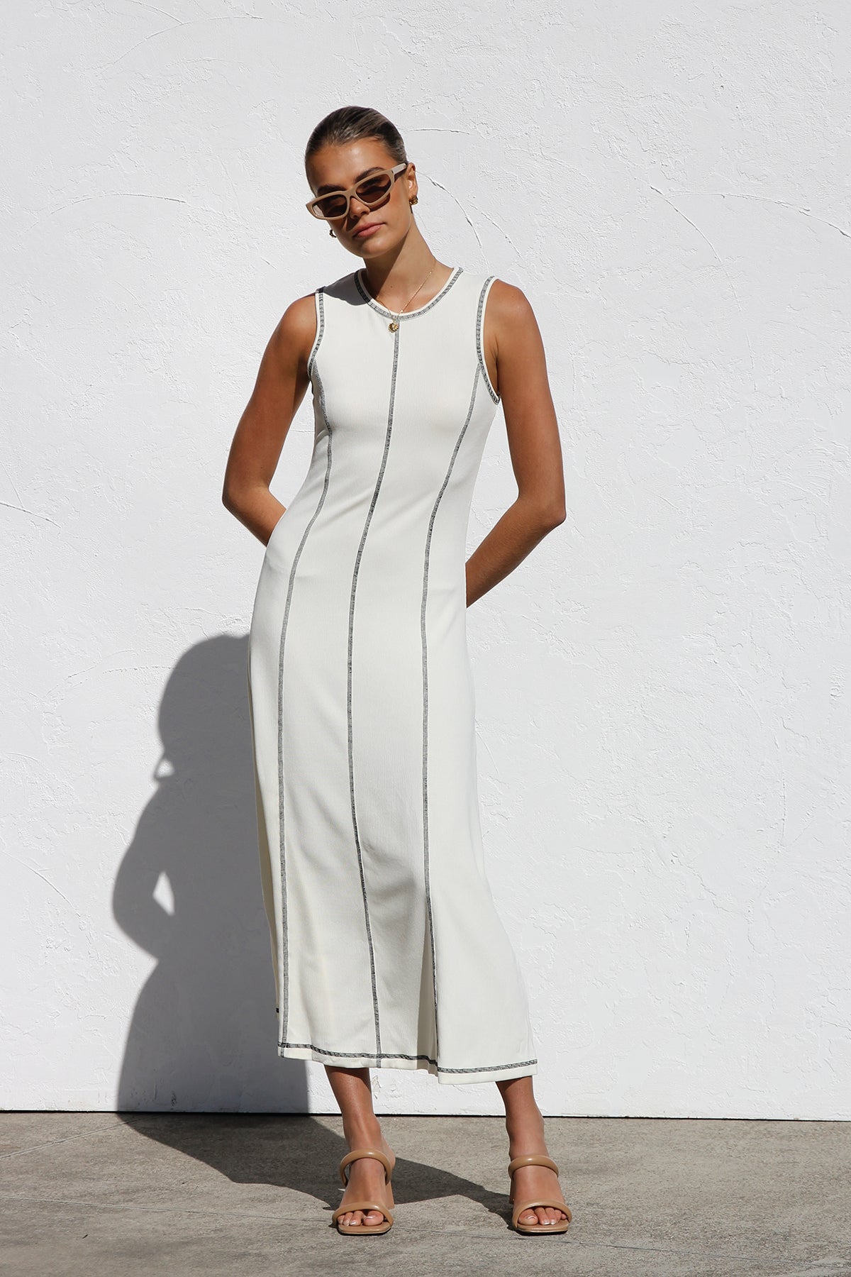 A woman stands against a white wall wearing a sleeveless, form-fitting, off-white midi dress. The dress has a high neckline and is detailed with dark vertical contrast stitching along the seams and armholes.
