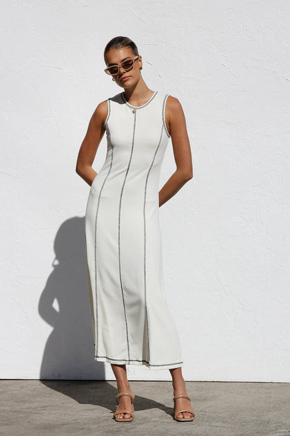 A woman stands against a white wall wearing a sleeveless, form-fitting, off-white midi dress. The dress has a high neckline and is detailed with dark vertical contrast stitching along the seams and armholes.