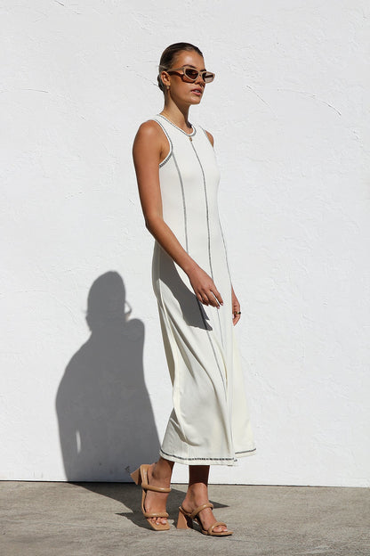 A woman stands against a white wall wearing a sleeveless, form-fitting, off-white midi dress. The dress has a high neckline and is detailed with dark vertical contrast stitching along the seams and armholes.