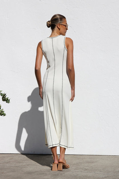 A woman stands against a white wall wearing a sleeveless, form-fitting, off-white midi dress. The dress has a high neckline and is detailed with dark vertical contrast stitching along the seams and armholes.