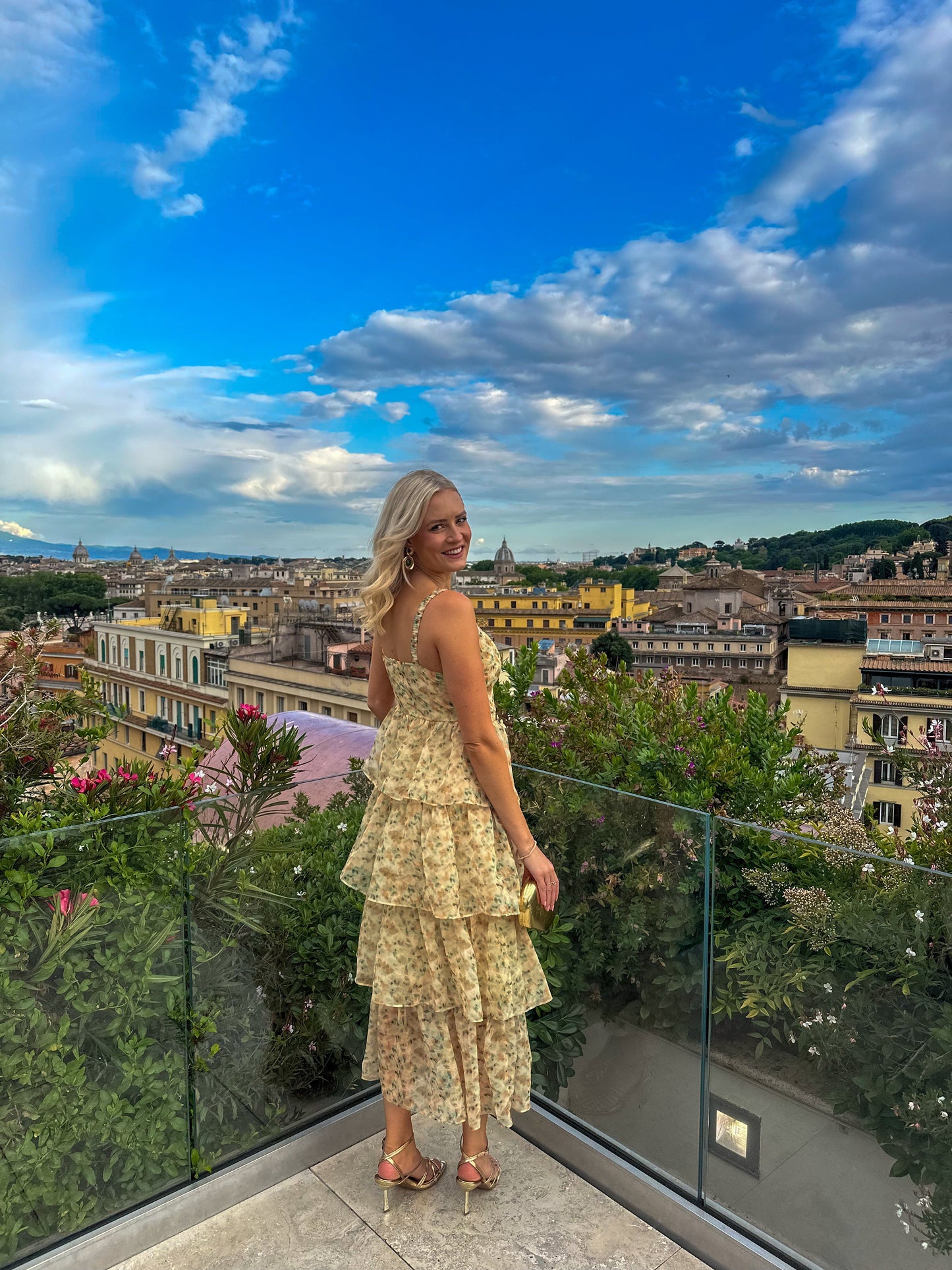 A woman stands on a scenic balcony wearing a romantic floral midi dress with a tiered ruffle skirt, sweetheart neckline, and a high leg slit.
