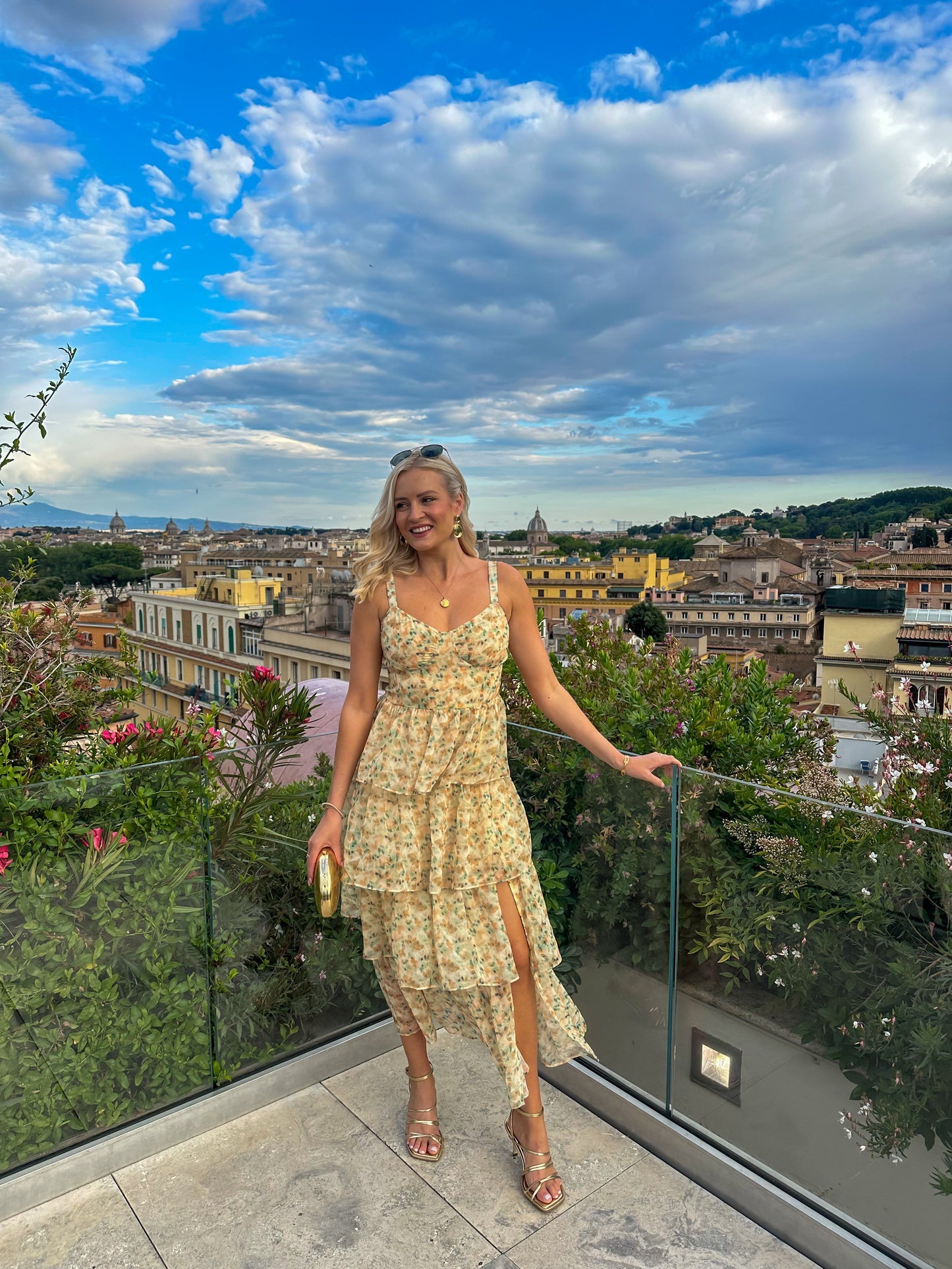 A woman stands on a scenic balcony wearing a romantic floral midi dress with a tiered ruffle skirt, sweetheart neckline, and a high leg slit.