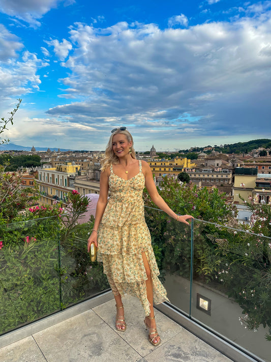 A woman stands on a scenic balcony wearing a romantic floral midi dress with a tiered ruffle skirt, sweetheart neckline, and a high leg slit.