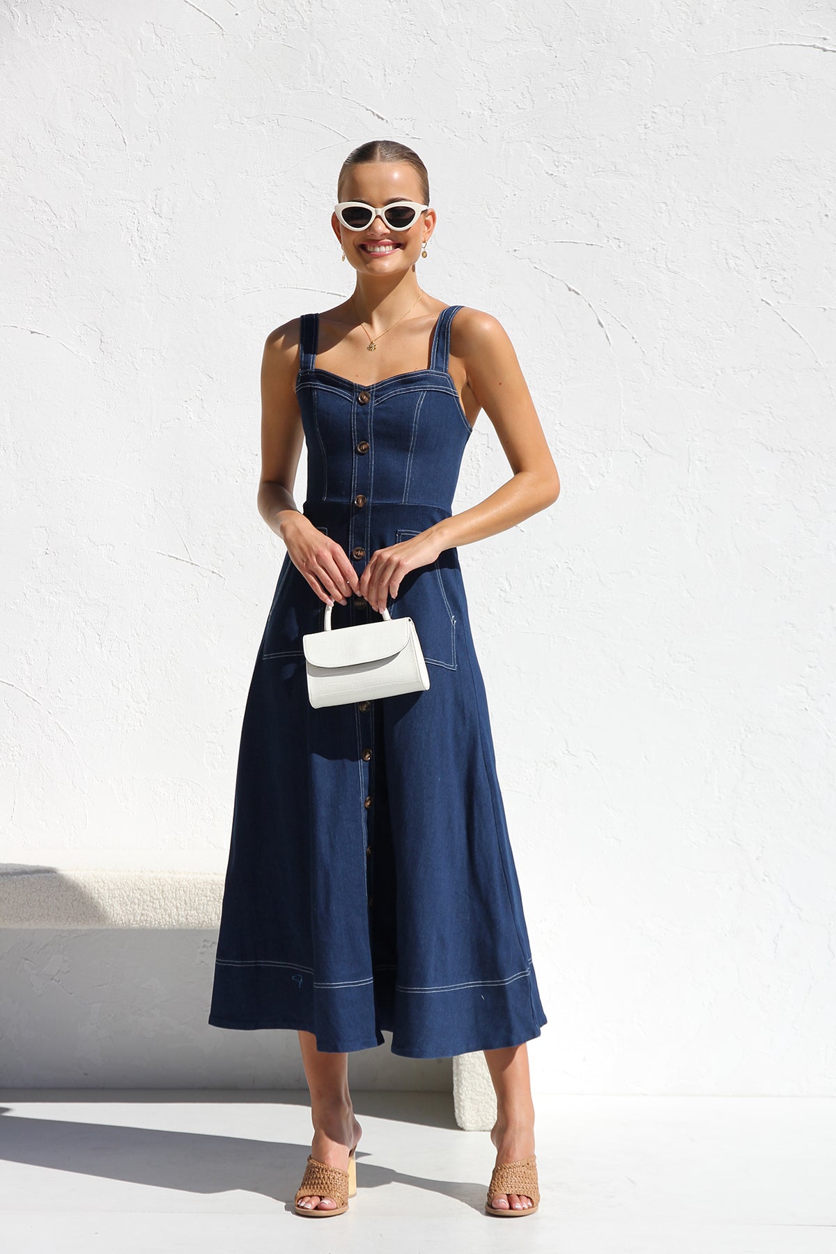 Indigo blue - A woman models a dark blue, sleeveless A-line midi dress with contrast white topstitching and a full button-front. She wears white cat-eye sunglasses and holds a small white handbag, standing against a white textured wall.
