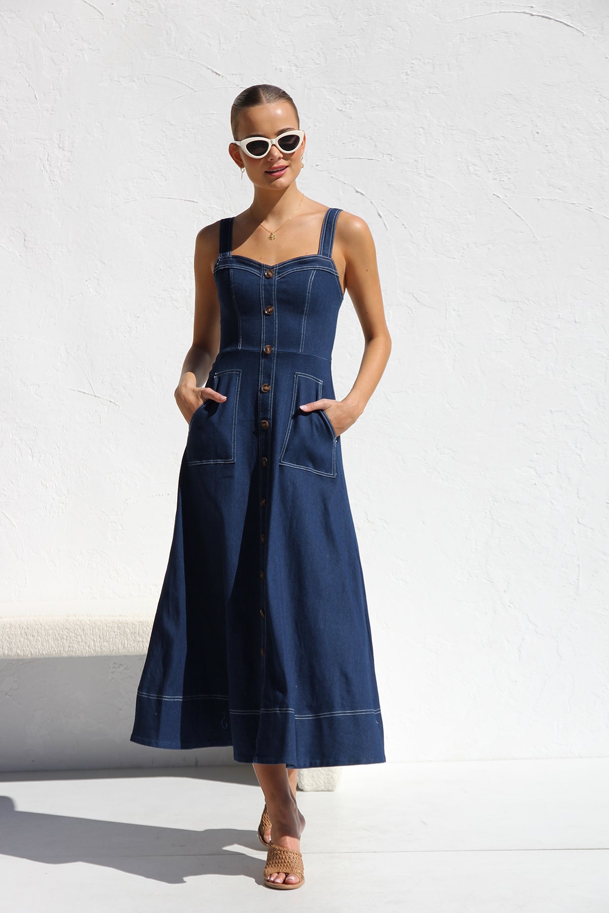 Indigo blue - A woman models a dark blue, sleeveless A-line midi dress with contrast white topstitching and a full button-front. She wears white cat-eye sunglasses and holds a small white handbag, standing against a white textured wall.