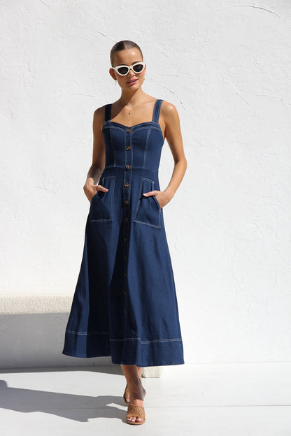 Indigo blue - A woman models a dark blue, sleeveless A-line midi dress with contrast white topstitching and a full button-front. She wears white cat-eye sunglasses and holds a small white handbag, standing against a white textured wall.