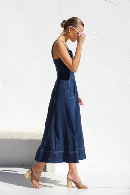 Indigo blue - A woman models a dark blue, sleeveless A-line midi dress with contrast white topstitching and a full button-front. She wears white cat-eye sunglasses and holds a small white handbag, standing against a white textured wall.