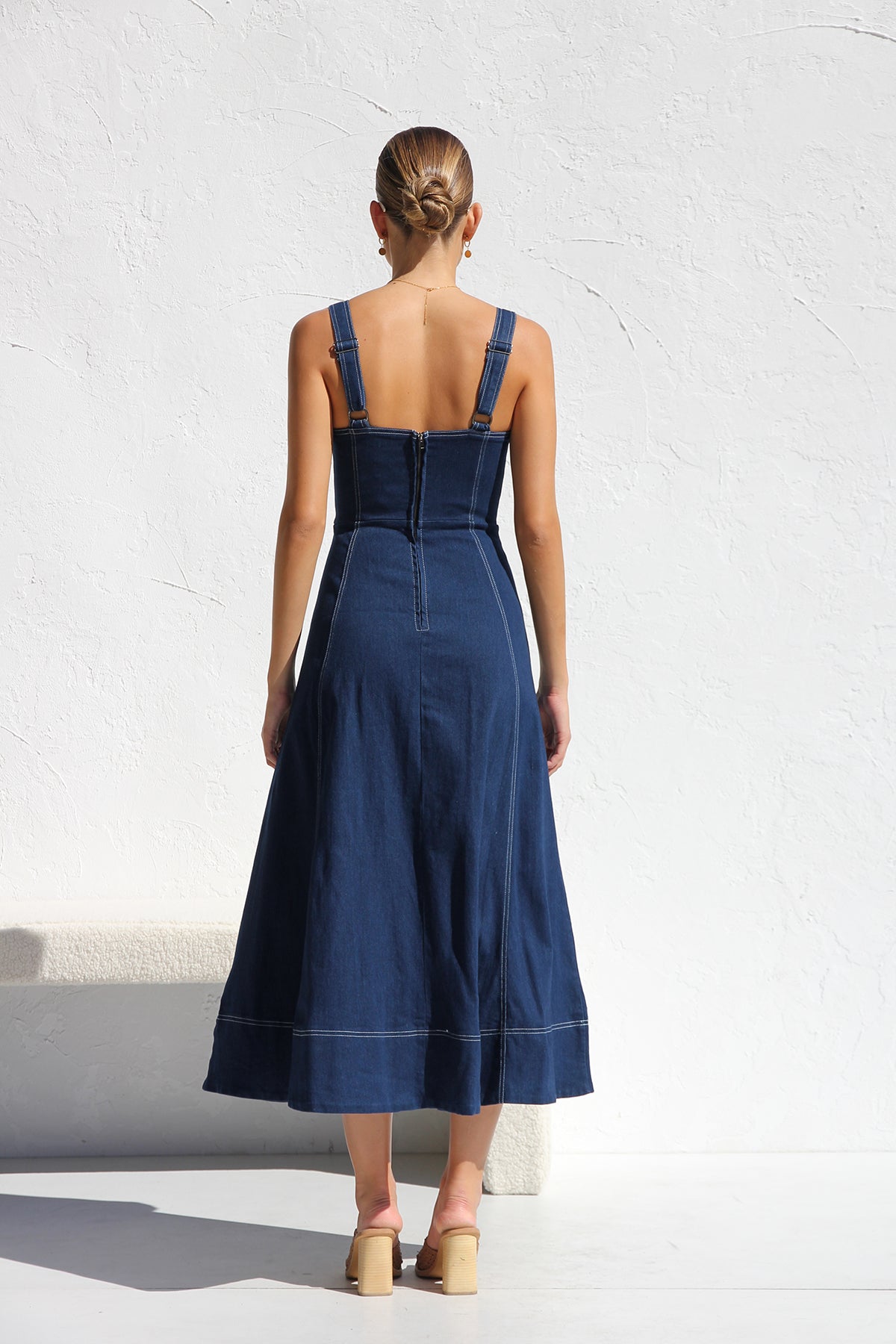 Indigo blue - A woman models a dark blue, sleeveless A-line midi dress with contrast white topstitching and a full button-front. She wears white cat-eye sunglasses and holds a small white handbag, standing against a white textured wall.
