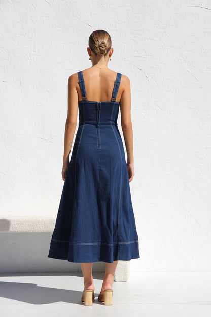 Indigo blue - A woman models a dark blue, sleeveless A-line midi dress with contrast white topstitching and a full button-front. She wears white cat-eye sunglasses and holds a small white handbag, standing against a white textured wall.
