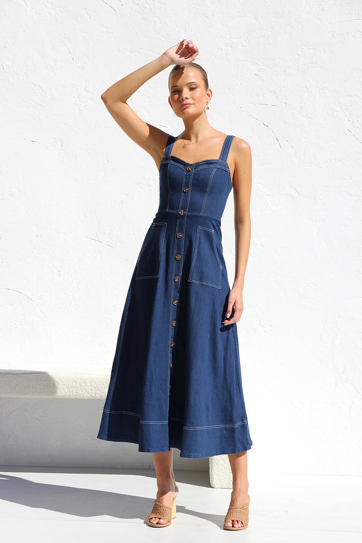 A woman models a dark blue, sleeveless A-line midi dress with contrast white topstitching and a full button-front. She wears white cat-eye sunglasses and holds a small white handbag, standing against a white textured wall.