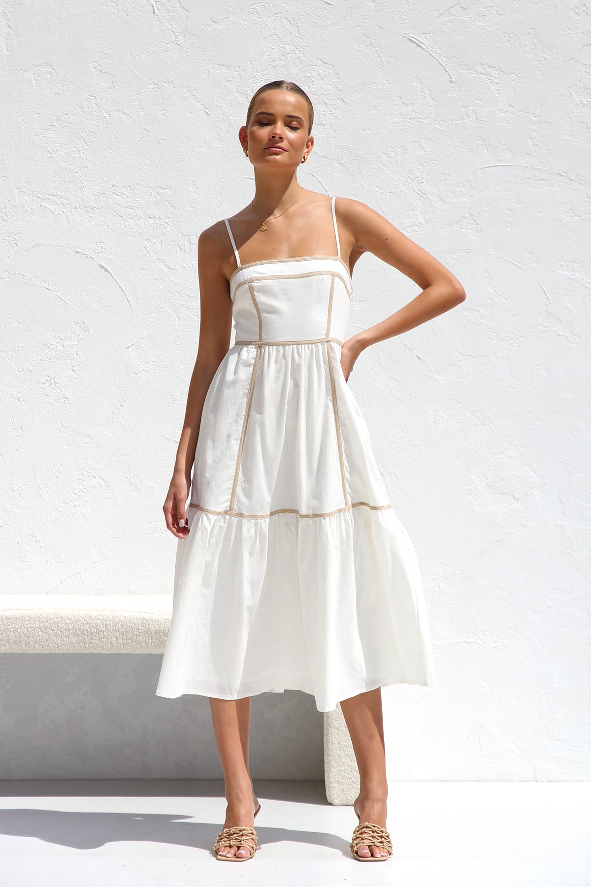 White - A woman wearing a white midi dress with thin straps and tan contrast piping on the bodice and tiered skirt. She is smiling and standing against a white textured wall, holding a woven clutch.