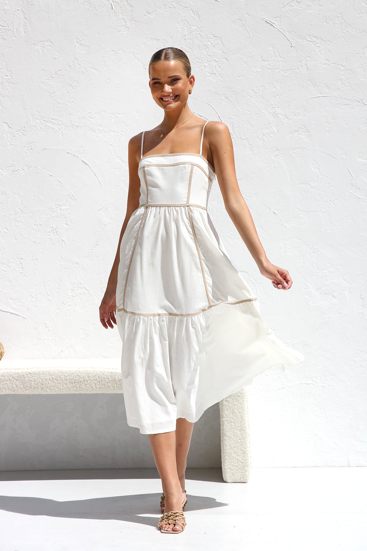 A woman wearing a white midi dress with thin straps and tan contrast piping on the bodice and tiered skirt. She is smiling and standing against a white textured wall, holding a woven clutch.