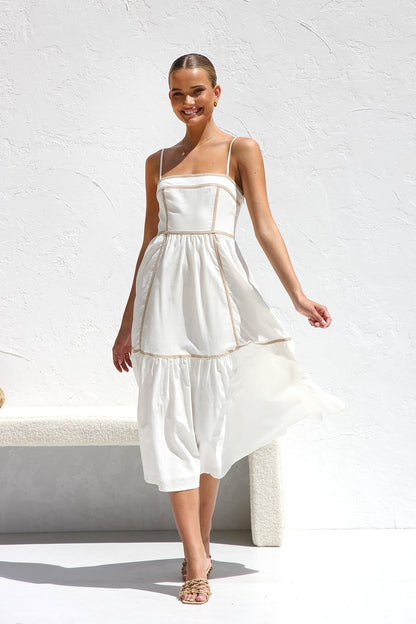 A woman wearing a white midi dress with thin straps and tan contrast piping on the bodice and tiered skirt. She is smiling and standing against a white textured wall, holding a woven clutch.