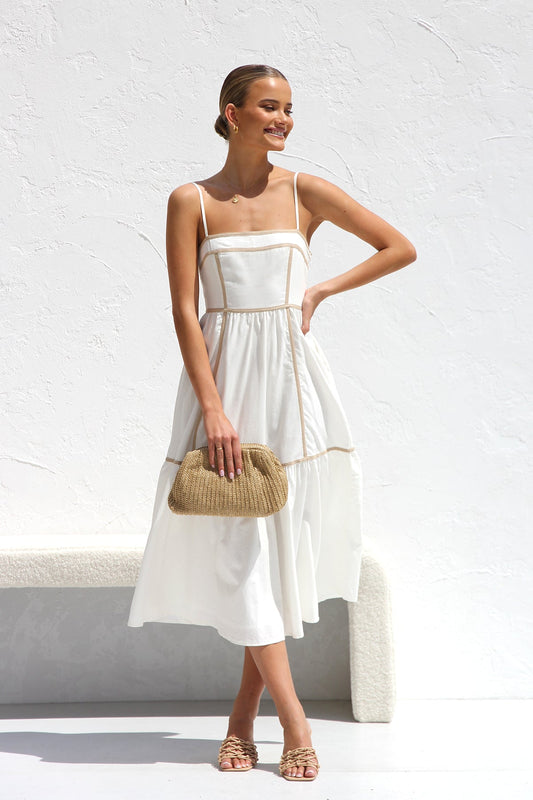 A woman wearing a white midi dress with thin straps and tan contrast piping on the bodice and tiered skirt. She is smiling and standing against a white textured wall, holding a woven clutch.