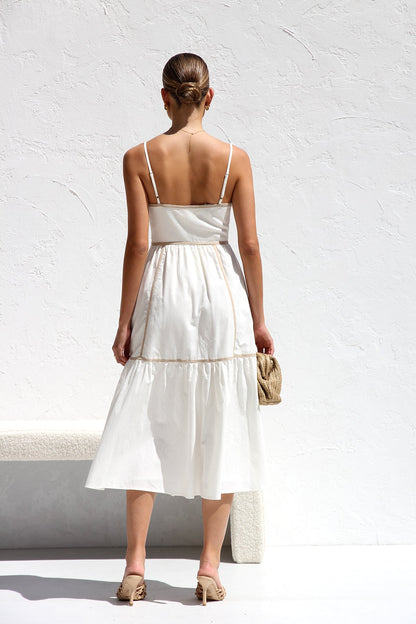 White - A woman wearing a white midi dress with thin straps and tan contrast piping on the bodice and tiered skirt. She is smiling and standing against a white textured wall, holding a woven clutch.