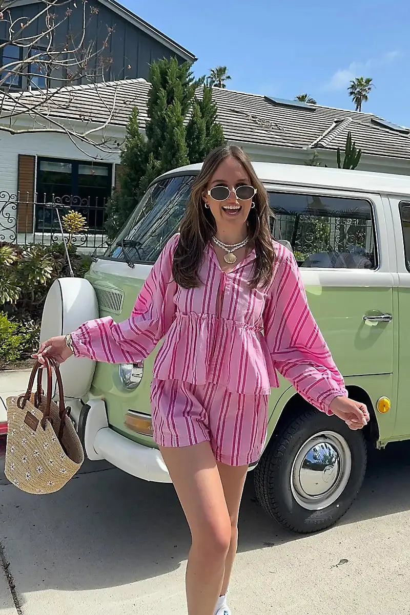 Pink - A woman wearing a relaxed-fit, green and white vertically striped two-piece set featuring a tiered peplum top with long balloon sleeves and a tie-front closure, paired with matching loose shorts, standing outdoors.