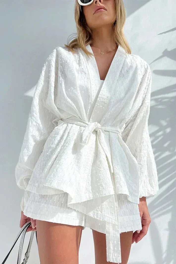 White - Woman wearing a white, lightweight wrap blouse featuring a textured dobby pattern, voluminous long sleeves, and a tie waist belt.