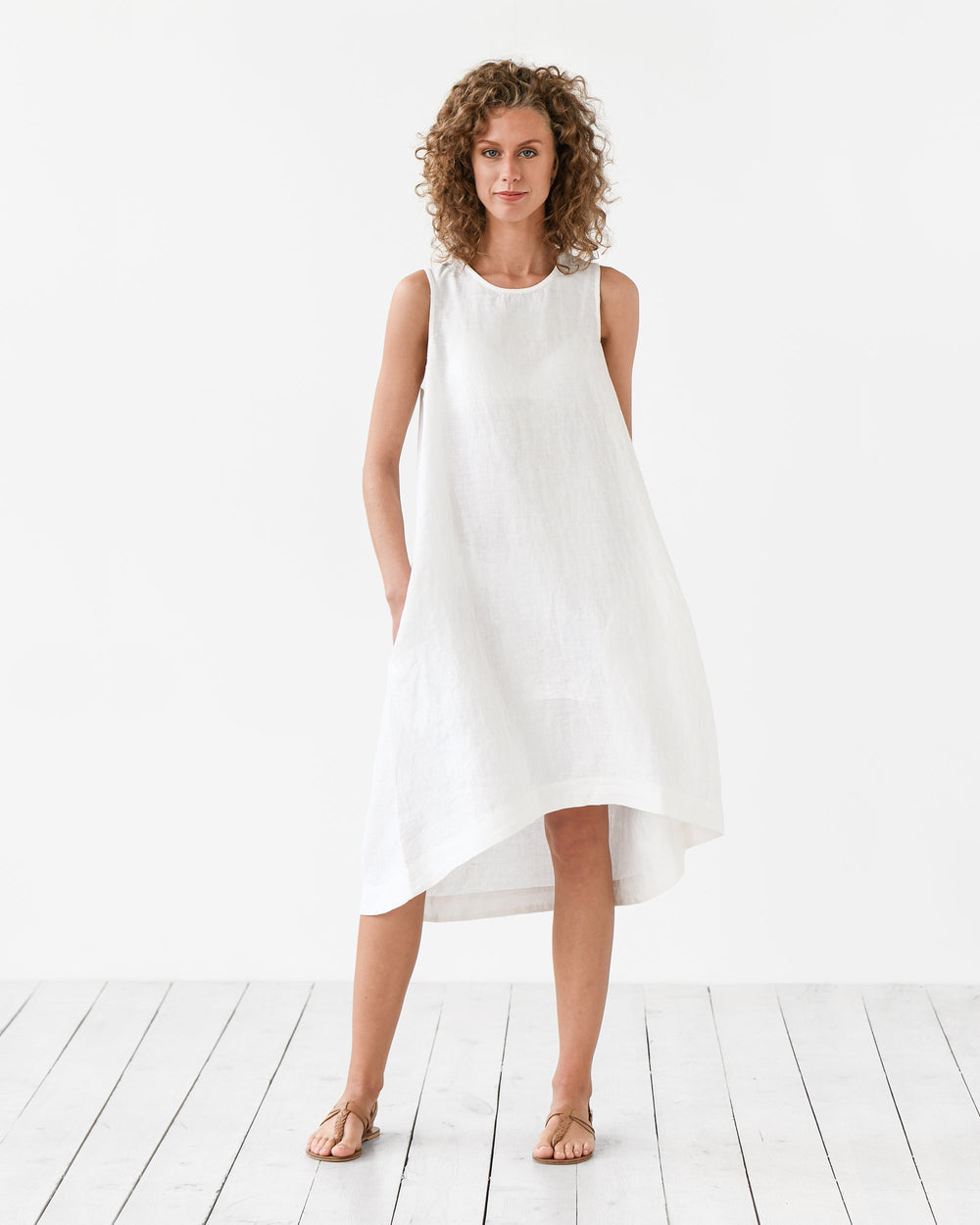 White - A woman wearing a relaxed, black sleeveless trapeze midi dress with a high-low hemline and round neckline, standing against a white textured wall.