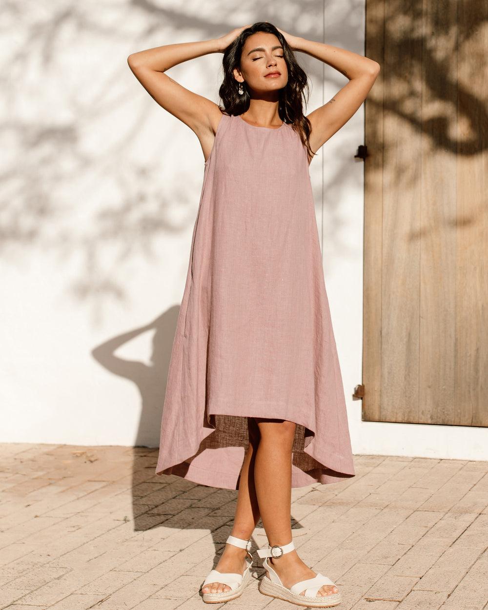 Rosewood - A woman wearing a relaxed, black sleeveless trapeze midi dress with a high-low hemline and round neckline, standing against a white textured wall.
