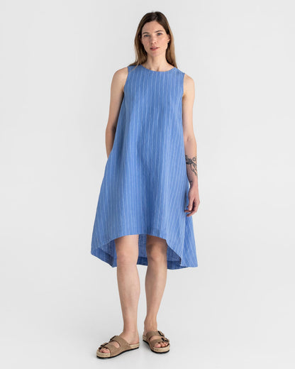 Blue - A woman wearing a relaxed, black sleeveless trapeze midi dress with a high-low hemline and round neckline, standing against a white textured wall.