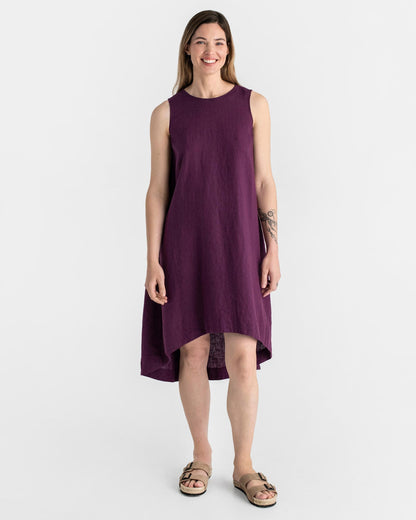 Purple - A woman wearing a relaxed, black sleeveless trapeze midi dress with a high-low hemline and round neckline, standing against a white textured wall.