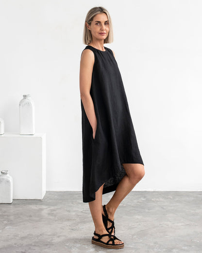 Purple - A woman wearing a relaxed, black sleeveless trapeze midi dress with a high-low hemline and round neckline, standing against a white textured wall.
