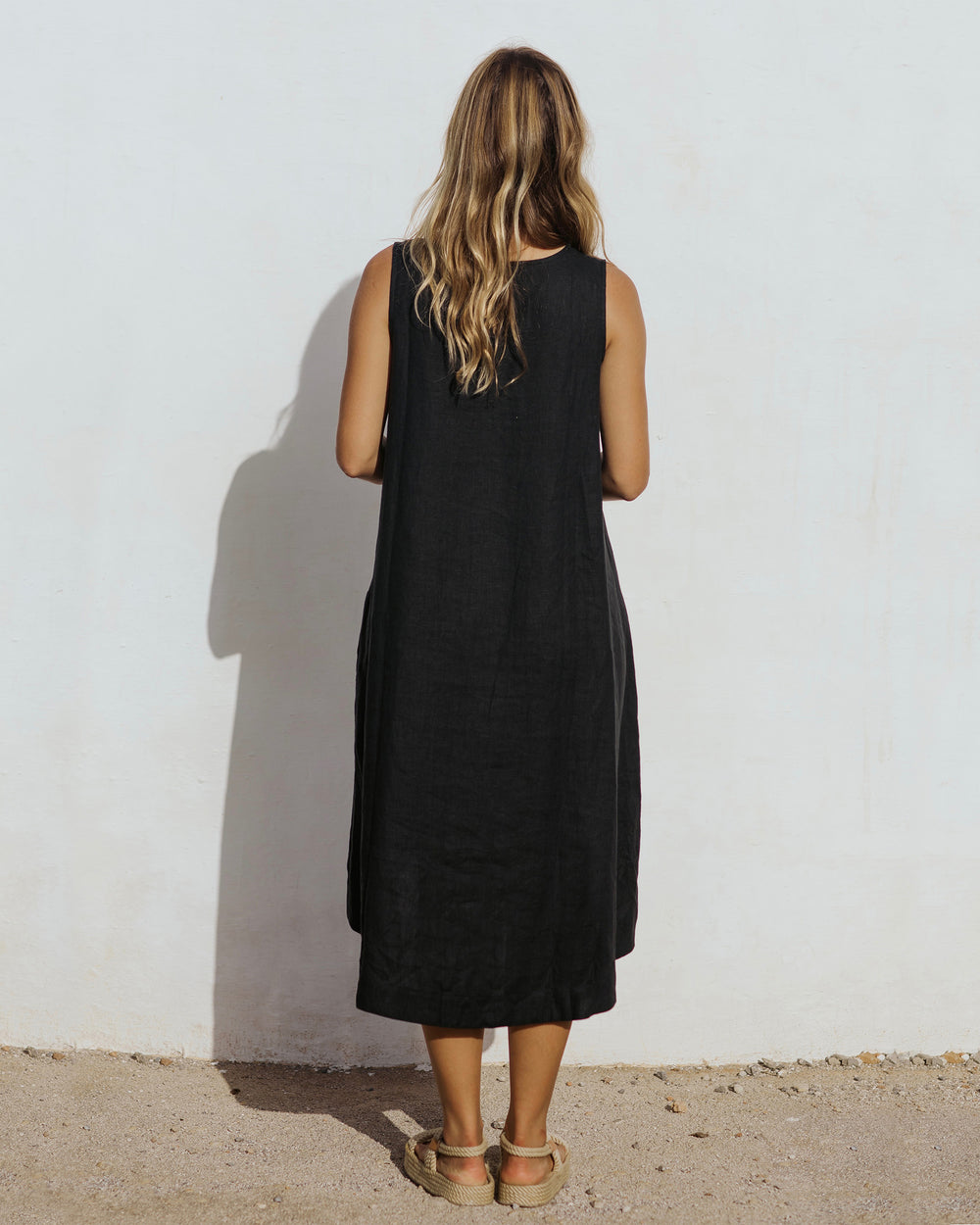 Purple - A woman wearing a relaxed, black sleeveless trapeze midi dress with a high-low hemline and round neckline, standing against a white textured wall.