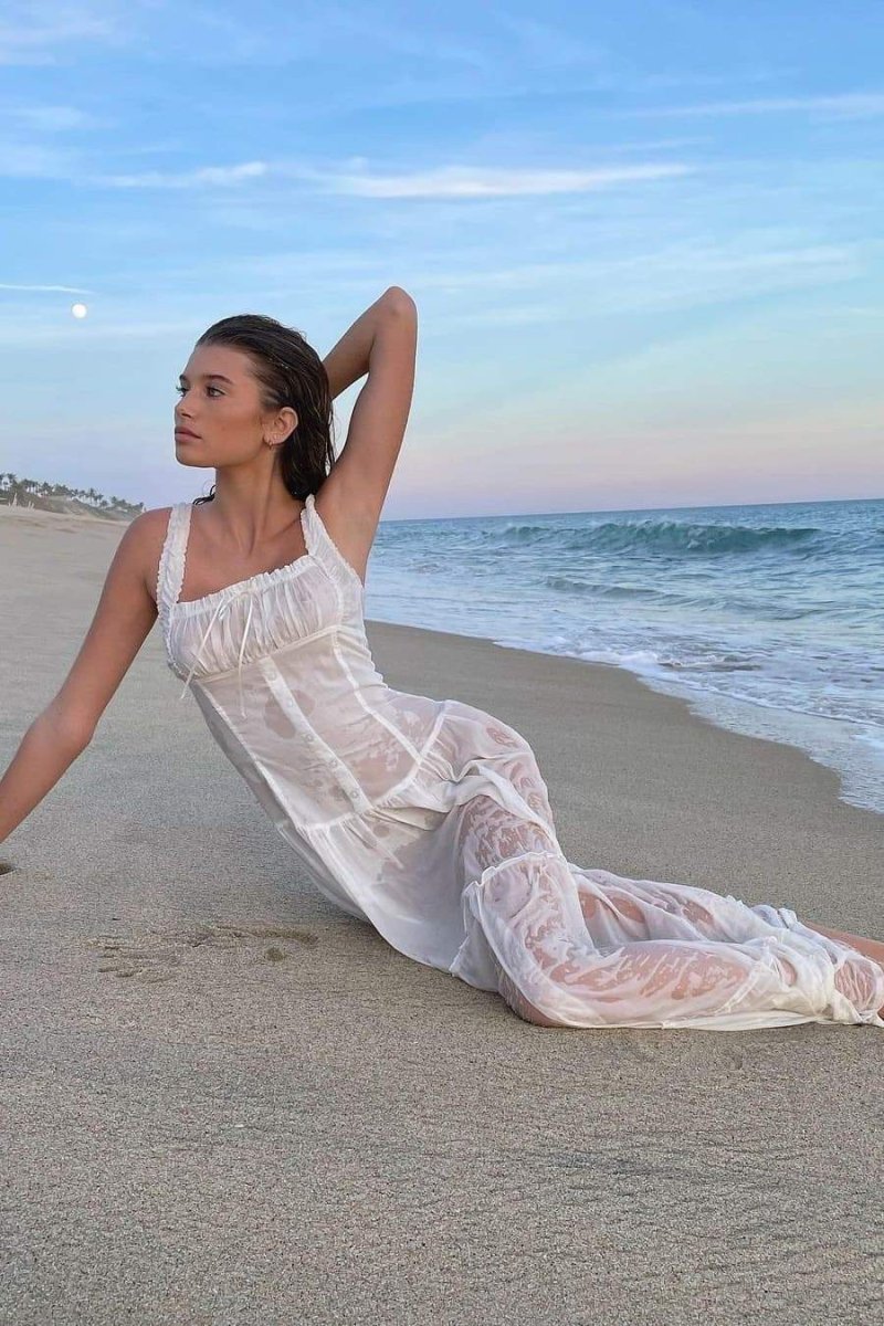 White - A woman sitting on rocks by the ocean wearing a white, sleeveless maxi dress featuring a ruched square neckline, corset-style buttoned bodice, and a voluminous tiered skirt.
