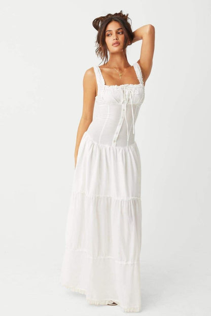 White - A woman sitting on rocks by the ocean wearing a white, sleeveless maxi dress featuring a ruched square neckline, corset-style buttoned bodice, and a voluminous tiered skirt.
