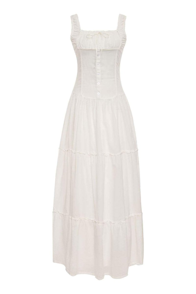 White - A woman sitting on rocks by the ocean wearing a white, sleeveless maxi dress featuring a ruched square neckline, corset-style buttoned bodice, and a voluminous tiered skirt.