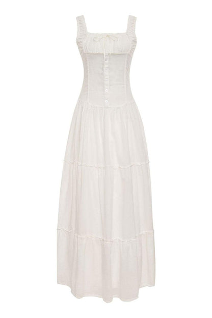 White - A woman sitting on rocks by the ocean wearing a white, sleeveless maxi dress featuring a ruched square neckline, corset-style buttoned bodice, and a voluminous tiered skirt.